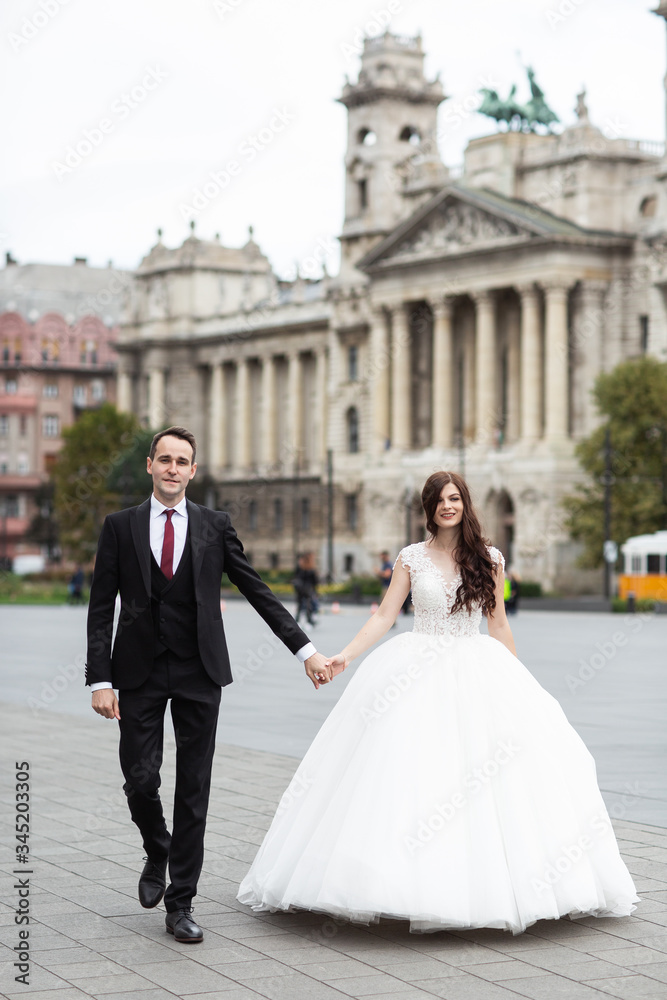 Fototapeta premium Bride and groom dancing in the old town street. Wedding couple walks in Budapest near Parliament House. Caucasian happy romantic young couple celebrating their marriage. Wedding and love concept.