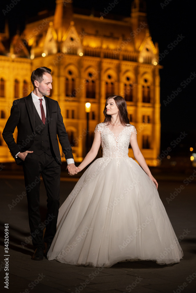 Naklejka premium Bride and groom hugging in the old town street. Wedding couple walks in Budapest near Parliament House in night. Happy romantic young couple celebrating their marriage. Wedding and love concept.