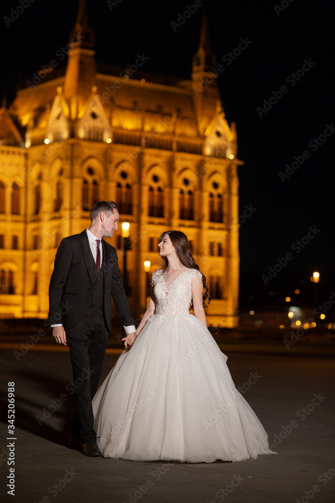 Bride and groom hugging in the old town street. Wedding couple walks in Budapest near Parliament House in night. Happy romantic young couple celebrating their marriage. Wedding and love concept.