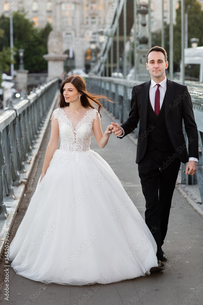 Bride and groom hugging in the old town street. Wedding couple walking on Szechenyi Chain Bridge, Hungary. Happy romantic young couple celebrating their marriage. Wedding and love concept.