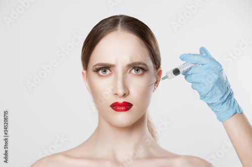 Portrait worried beautiful woman afraid of anti aging serum shot in cheekbones plastic surgery doctor's hand blue gloves holding injection over woman's face isolated white wall. Negative expression