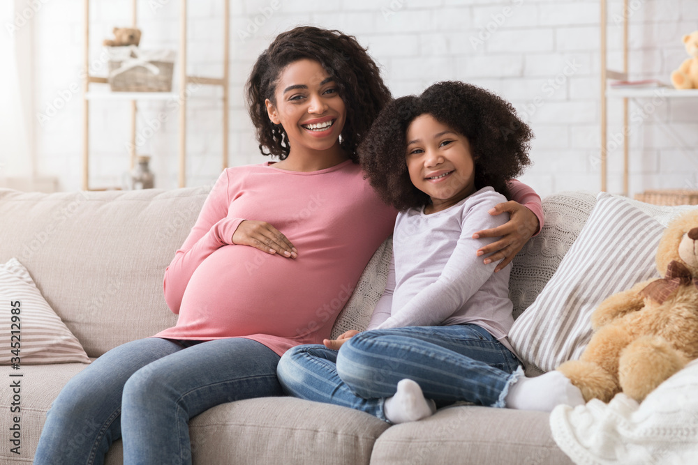 Smiling black pregnant woman and her cute little daughter posing at home