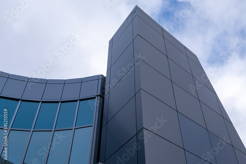 The corner of a grey coloured building made of metal composite panels and green reflective windows. Part of the building has a high tower and a lower curved section made of green windows. 