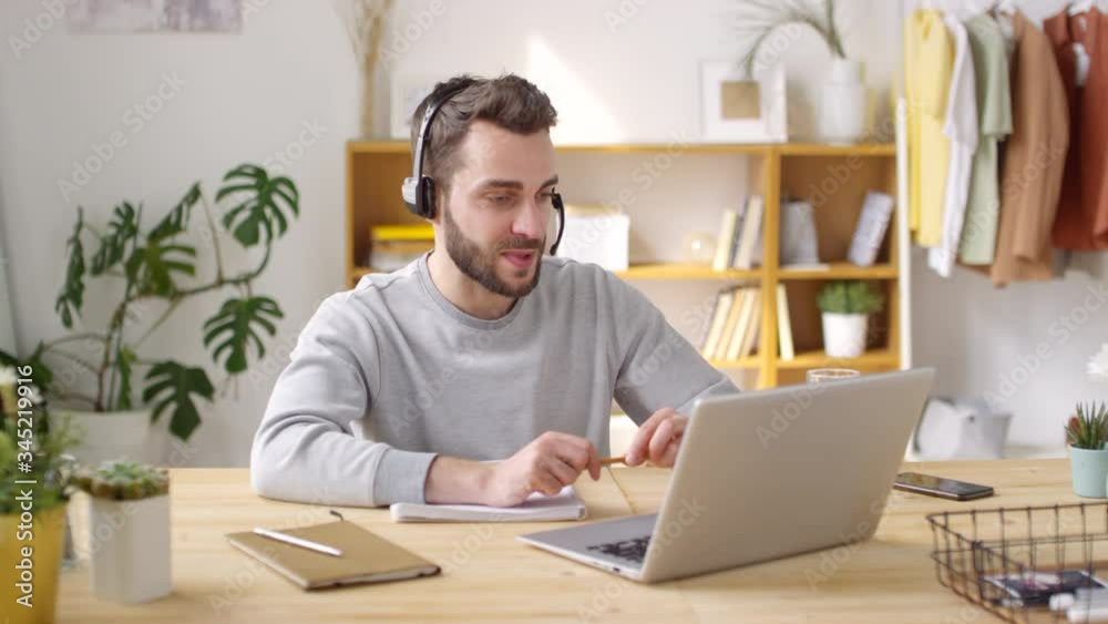 Medium shot of cheerful young man in wireless headset talking on video call on laptop while working remotely from home, then ending call and smiling for camera