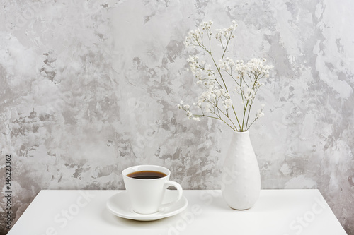 Cup of coffee and bouquet of small delicate flowers gypsophila in vase on white table agains gray stone wall. Copy space Minimal style. Concept Good morning and coffee time