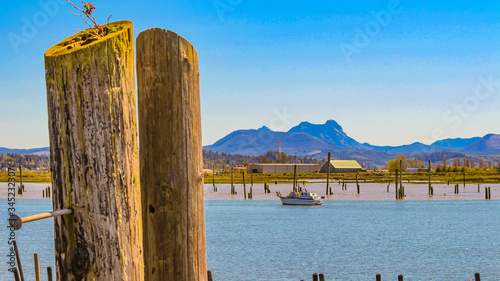 A fishing boat at idle work, with Saddle mountain in the background. Two old Pilings in the forefront one with a large nail protruding. In the fishing port of Astoria, Oregon.