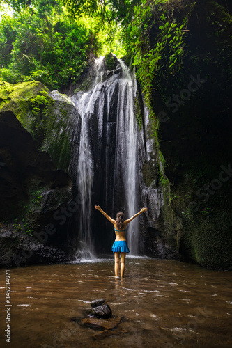 Excited Caucasian woman raising arms in front of the waterfall. Travel concept. View from back. Krisik waterfall. Bangli, Bali, Indonesia