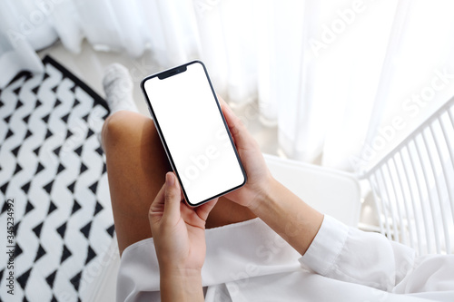 Top view mockup image of woman holding mobile phone with blank desktop white screen while sitting in bedroom with feeling relaxed