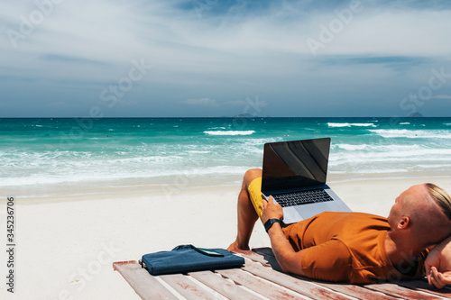 Young guy with long hair by the sea on a sunny day, lies with a laptop on her lap, working remotely with a business. Blogger, freelancer, Internet, work travel. Summer vacation