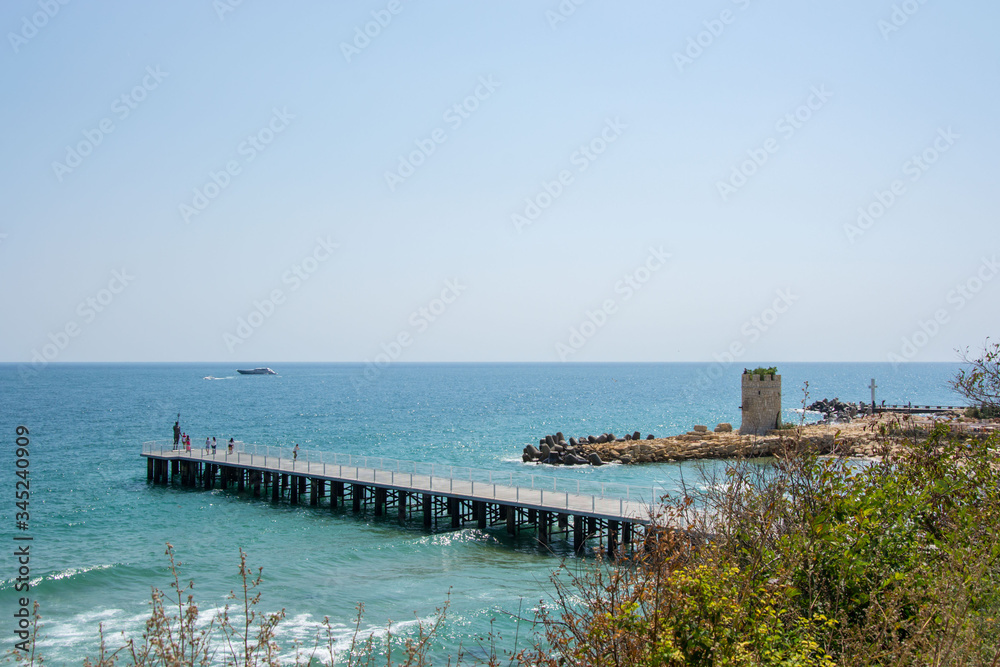 People walking on a pier with statue of Poseidon, bridge near the beach, blue sea and sky