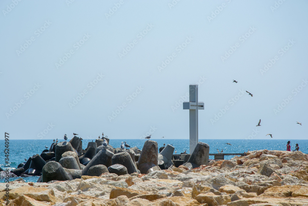 Seascape, view of a big concrete cross on the bay, people walking and birds flying, blue waters and sky, breakwaters, summer season, nature