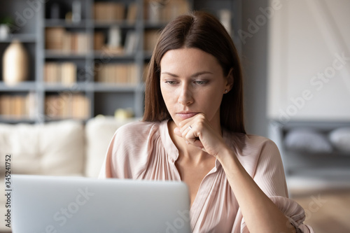 Thoughtful woman looking at laptop screen, pondering project, working online at home, sitting at desk, pensive young female touching chin, reading email, financial report, making decision
