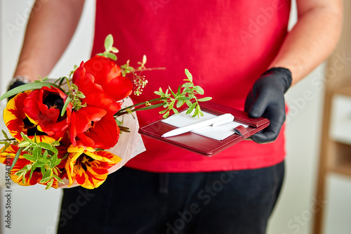 Courier, delivery man in red in medical latex gloves safely delivers online purchases a bouquet of flower during coronavirus epidemic. Stay home, safe concept. Contactless delivery service under quara photo