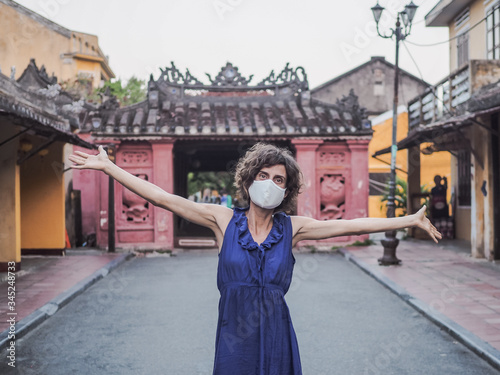 women wearing a face mask protecting from corona virus covid-19 standing in empty street arms up in front of japanese covered bridge in hoi an vietnam 