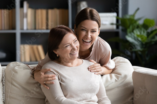 Excited laughing young woman hugging smiling older mother from back, family having fun, adult daughter and mature mom spending leisure time together at home, two generations bonding photo