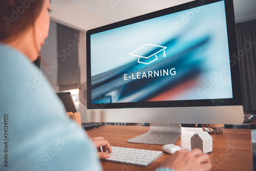 Online education, e-learning. Young woman is sitting at table, working on computer monitor with inscription on screen e-learning and image of square academic cap, distance training.