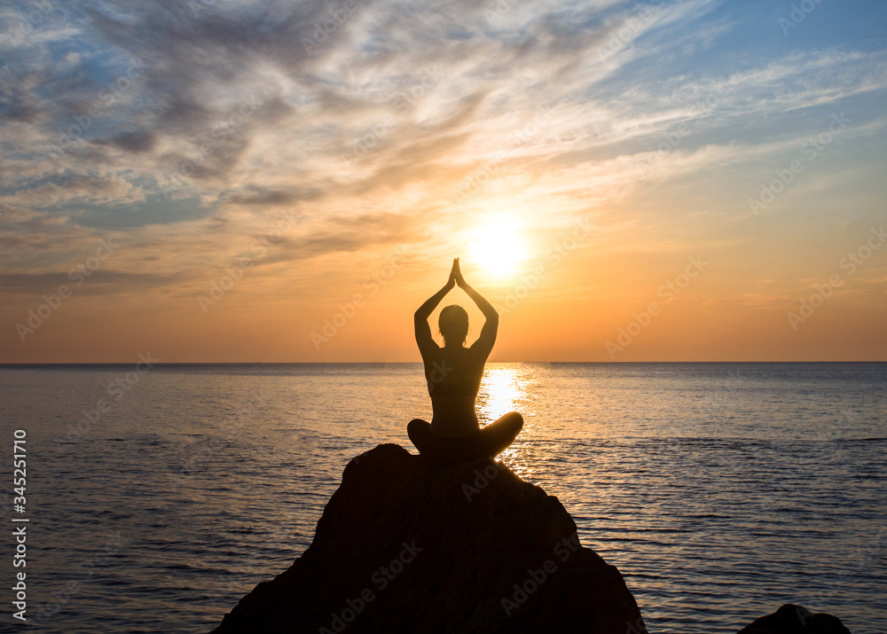 girl is sitting in lotus pose on the big rock in front of sunrise at the sea