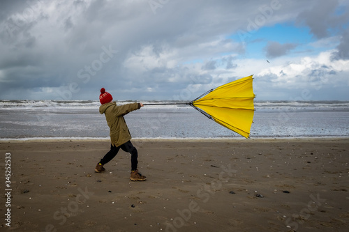 Young boy struggling with large yellow umbrella on beach in stormy weather. Winter beach scene.