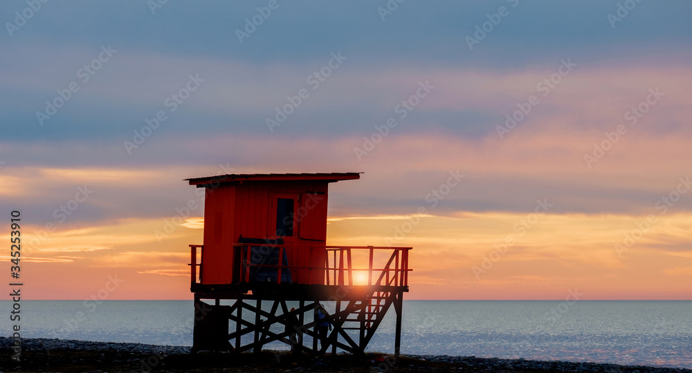 Red wooden lifeguard rescue tower on the empty beach with sunset cloudy sky background