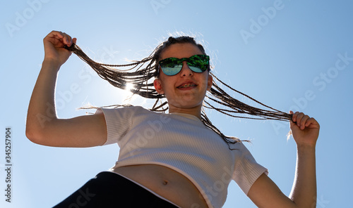 White teenager with hair braids wearing a white top and sunglasses singing a song with the sky at the background. The teenager is stretching her braids. Horizontal photo