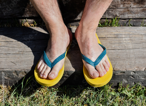 a couple of feet completely barefoot wearing two yellow and blue flip flops on a wooden step. The man is resting in the garden in a sunny day of summer. Horizontal photo .