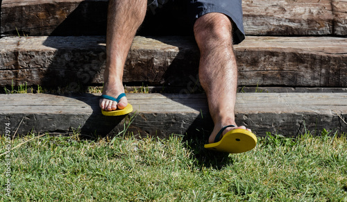 a couple of feet completely barefoot wearing two yellow and blue flip flops. The man wearing blue shorts is sat on a step of the wooden stairs in the garden in a summer sunny day. Horizontal photo .