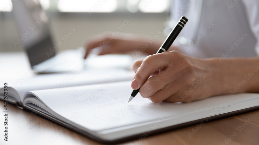 Female professional doctor hand making notes in medical journal using laptop computer sitting at desk. Woman physician, nurse or pharmacist wearing white coat writing in paper notebook. Close up view Female professional doctor hand making notes in medical journal using laptop computer sitting at desk. Woman physician, nurse or pharmacist wearing white coat writing in paper notebook. Close up view