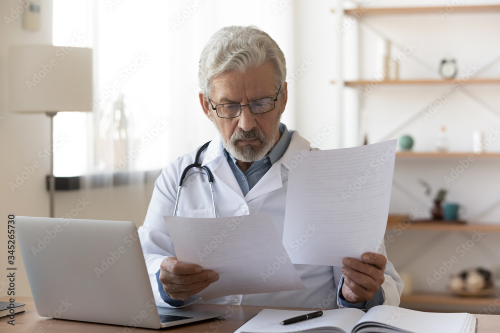 Serious professional senior elderly doctor doing paperwork checking medical documents at workplace. Concentrated old physician reading medic form analyzing patient diagnosis or report in hospital.