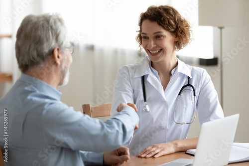 Smiling young female doctor handshaking greeting healthy senior male patient in hospital. Happy practitioner shake hand of elder client at advice meeting. Old people medical health care trust concept.