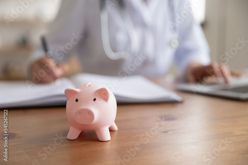 Female doctor working at desk with piggy bank box. Piggybank on table as concept of healthcare fees financial cost, money savings on health care insurance, medical care expenses concept. Close up view photo