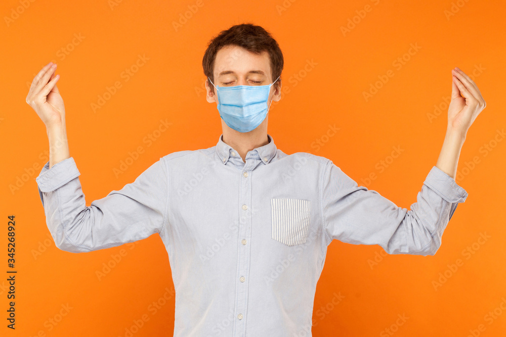 Yoga and meditation. Portrait of calm young worker man with surgical medical mask standing with closed eyes and meditating. indoor studio shot isolated on orange background. Yoga and meditation. Portrait of calm young worker man with surgical medical mask standing with closed eyes and meditating. indoor studio shot isolated on orange background.