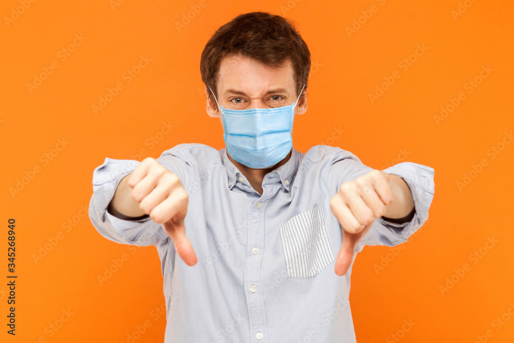 I don't like it. Portrait of sad young worker man with surgical medical mask standing thumbs down and looking at camera with dissatisfied face. indoor studio shot isolated on orange background. I don't like it. Portrait of sad young worker man with surgical medical mask standing thumbs down and looking at camera with dissatisfied face. indoor studio shot isolated on orange background.