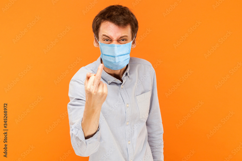 Portrait of angry young worker man with surgical medical mask standing looking at camera with middle finger and aggressive face. indoor studio shot isolated on orange background. Portrait of angry young worker man with surgical medical mask standing looking at camera with middle finger and aggressive face. indoor studio shot isolated on orange background.