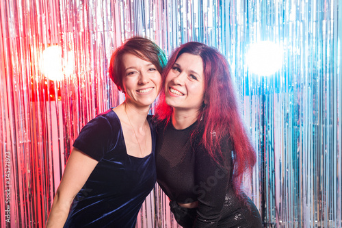 Portrait of two attractive happy women in evening dresses hugging and looking at camera isolated over silver coloured shiny background. Birthday party, holidays and night club concept.