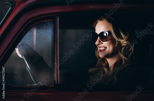 Blonde caucasian woman is smiling and driving retro red car at night. Film looking photo of girl driving in enjoyment. James Bond style photo.
