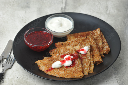 On a black plate, thin pancakes with sour cream and jam.  Light background.  Close-up.  In the frame are cutlery.  