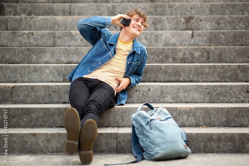 Full body happy young man relaxing on steps outside talking with cellphone and looking away