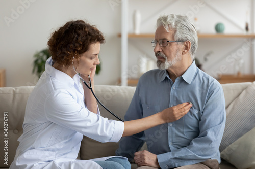 Female attending physician holding stethoscope listening old patient during homecare visit. Doctor checking heartbeat examining elder retired man at home. Seniors heart diseases, cardiology concept.