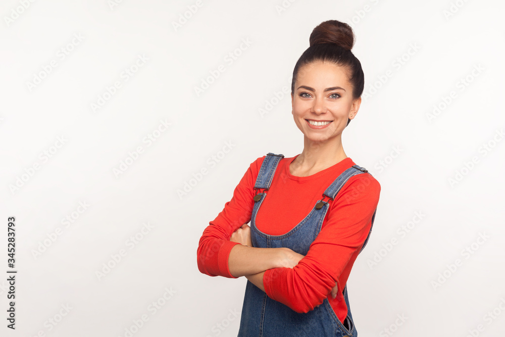 Portrait of cheerful happy stylish pretty girl with hair bun in denim overalls standing with crossed hands and smiling, expressing confidence, optimism. indoor studio shot isolated on white background Portrait of cheerful happy stylish pretty girl with hair bun in denim overalls standing with crossed hands and smiling, expressing confidence, optimism. indoor studio shot isolated on white background