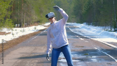 Woman in cap dancing in forest on country road uses vr googles waving hands. Playing games or watching 3d films on virtual reality headset in Augmented Reality. Challenge of dance on street in spring.