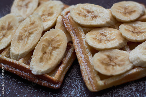 Belgian waffles with banana and sugar powder on dark background.