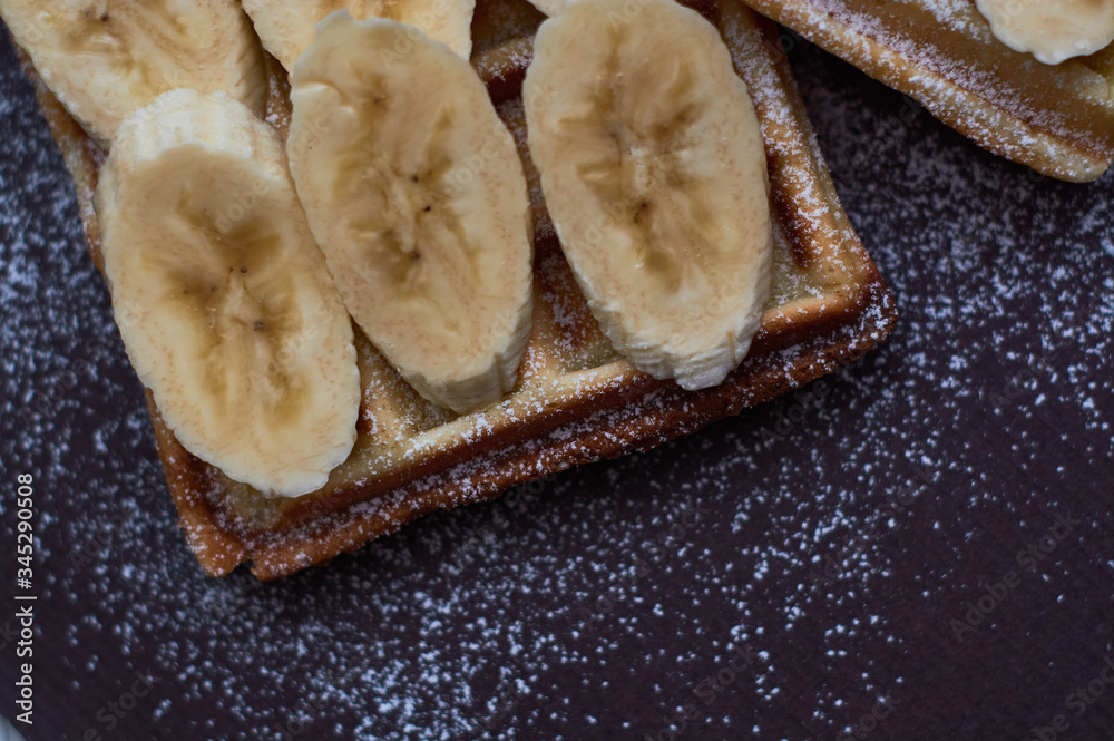 Belgian waffles with banana and sugar powder on dark background.