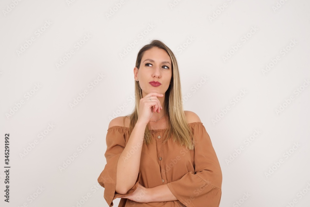 Shot of contemplative thoughtful Young woman keeps hand under chin, looks thoughtfully upwards, dressed in casual clothes, poses over white background with free space for your text Shot of contemplative thoughtful Young woman keeps hand under chin, looks thoughtfully upwards, dressed in casual clothes, poses over white background with free space for your text