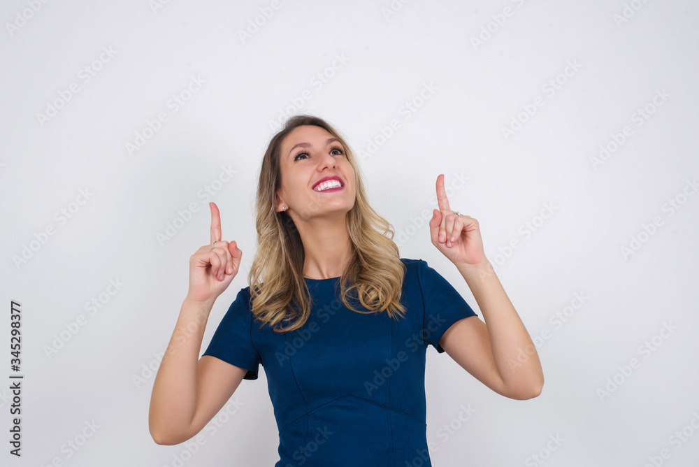 Horizontal shot of successful friendly looking young dark haired woman with bright make up exclaiming excitedly, pointing both index fingers up, indicating something shocking on blank copy space wall. Horizontal shot of successful friendly looking young dark haired woman with bright make up exclaiming excitedly, pointing both index fingers up, indicating something shocking on blank copy space wall.