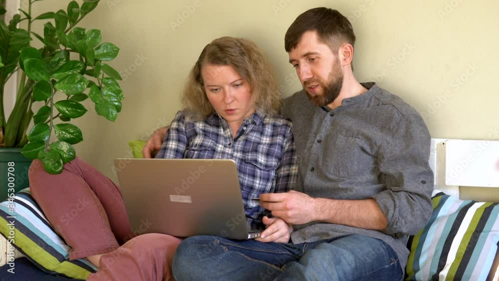 Young Caucasian couple doing shoppings online on the couch at home with laptop, looking at each other and smiling. Selecting goods from Internet store. Young Caucasian couple doing shoppings online on the couch at home with laptop, looking at each other and smiling. Selecting goods from Internet store.