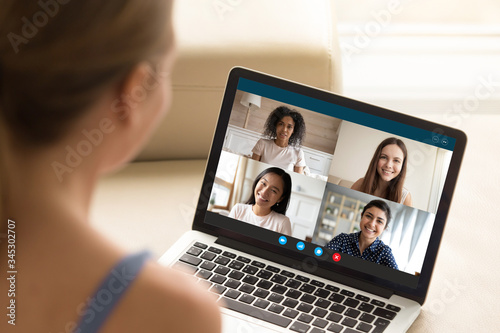 Back view of millennial girl talk chat on video call on laptop with smiling diverse multiethnic girlfriends, young woman have fun engaged in webcam conference on computer with female friends
