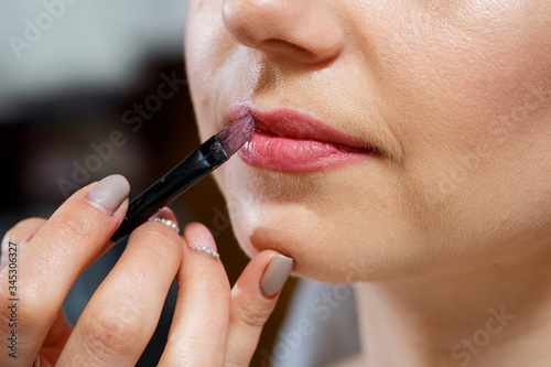 Makeup artist applies red lipstick. Beautiful female face. Hand of a make-up master painting lips of a young girl beauty model. Makeup in progress