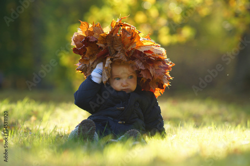 Little thoughtful child with yellow wreath of maple leaves on head sitting on grass in sunny autumn park