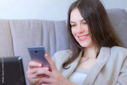 Woman working typing on laptop and reading message on smartphone sitting on couch smiling, distant work. Coronavirus pandemic and quarantine at home. Self empoyed businesswoman, freelancer. photo