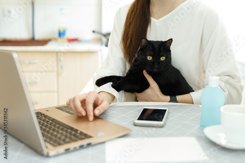 woman girl in a medical mask works on a laptop, holds a black cat in her arms, social distance during covid-19